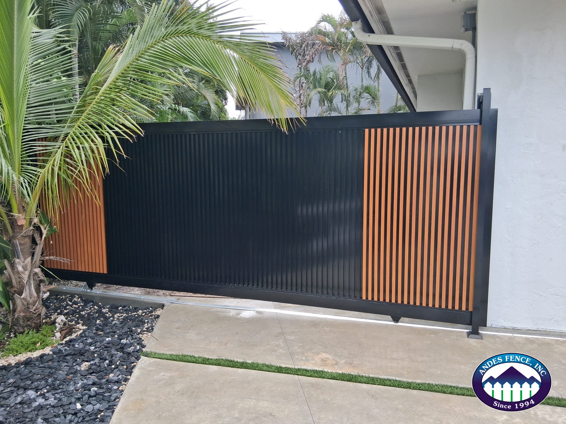 Black sliding gate with vertical slats and wood-look aluminum accents beside a home