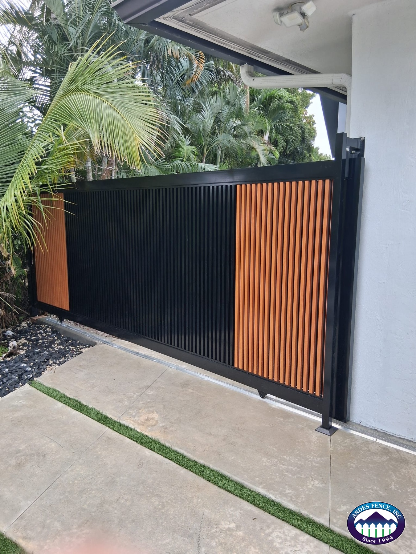 Angled view of a black vertical-slat sliding gate with wood-look aluminum accents and a ground track
